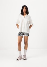 Camicetta bianca a maniche corte con taschino, abbinata a pantaloni corti a fantasia in bianco e nero, scarpe da ginnastica rosa e calze lunghe grigie chiare.