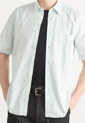 Person trägt ein hellmintfarbenes kurzärmeliges Hemd über einem schwarzen T-Shirt, dazu dunkle Jeans und einen schwarzen Gürtel, die Hände in den Taschen.