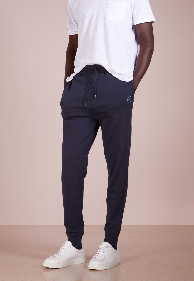 Marina blå bomullsbyxor med dragsko i midjan, ribbade manschetter och ett litet logopatch på fickan, kombinerat med vita sneakers.