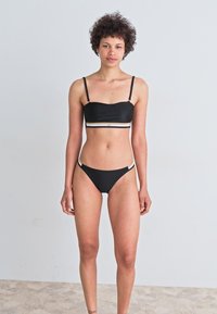 Μαύρο σετ μπικίνι με αμάνικο τοπ με λεπτές τιράντες και σλιπ με χαμηλή ίσια κοπή. Διακοσμημένο με άσπρη και μπεζ λωρίδα. Λείος υφή.
