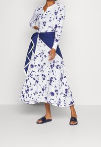 Robe à motifs floraux avec des manches longues, en blanc et bleu marine, dotée d'un détail enveloppant bleu marine et d'une silhouette fluide ; associée à des sandales bleu marine.