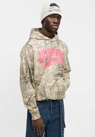 Φούτερ καμουφλάζ σε μπεζ με ροζ κείμενο "Billionaire Boys Club", διαθέτει τσέπη καγκουρό και κουκούλα με κορδόνι, συνδυασμένο με μπλε τζιν.