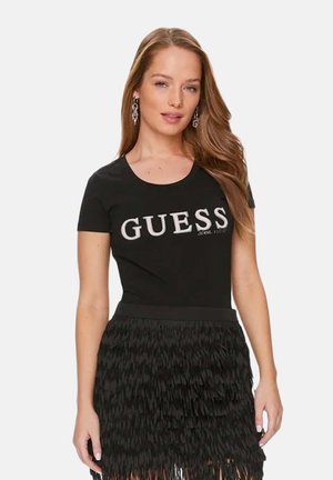 Femme aux longs cheveux portant des boucles d'oreilles argentées, un t-shirt noir Guess et une jupe noire texturée à franges, sur un fond uni.