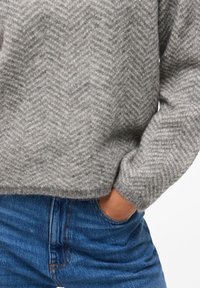 Suéter cinza com padrão de espinha de peixe, feito de material macio, com um corte descontraído e punhos canelados. Combinado com jeans azul de denim.