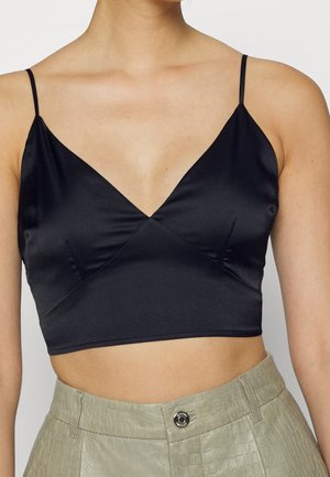 Kobieta nosząca czarny krótki satynowy top na cienkich ramiączkach w połączeniu z beżowymi, wysokim stanem, teksturowanymi spodniami.