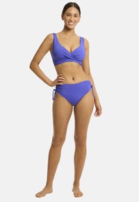 Model in een levendige paarse bikini, zelfverzekerd glimlachend, met één hand op haar heup en de andere ontspannen naast zich, tegen een effen achtergrond.