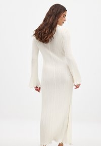 Lång, figurnära klänning i ribbat tyg, i off-white färg, med långa ärmar med utsvängda manschetter och en scallopad nederdel.
