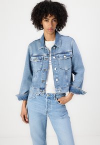Giacca di jeans blu chiaro con un design accorciato, dotata di tasche frontali, chiusura a bottoni e cuciture a contrasto. Indossata sopra una maglietta bianca.