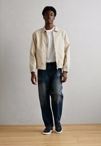 Beige lett jakke, hvit t-skjorte, mørkeblå jeans og mørke sko med hvite såler. Enkelt design med avslappet passform og glatt tekstur.