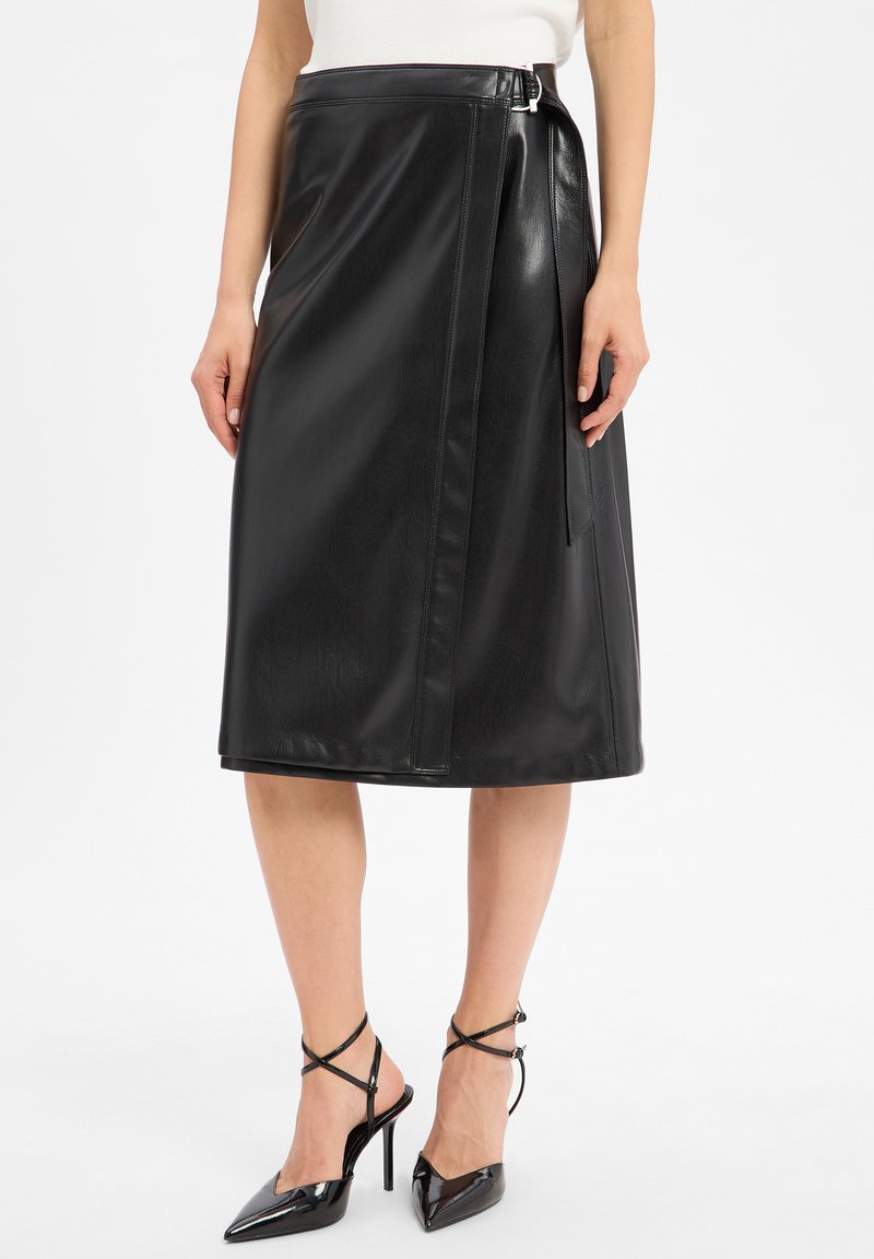 Jupe midi en simili cuir noir avec un design enveloppant, un détail de ceinture et une texture lisse. Assortie à des escarpins noirs à bout pointu et à des brides croisées.