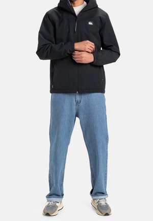 Osoba v černé kapuci, světle modrých džínách a šedých teniskách stojící s rukama sepnutýma před sebou na jednobarevném pozadí.