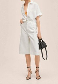 Chemise blanche à manches courtes avec boutons, associée à un pantalon culotte blanc taille haute. Sandales noires à brides croisées et petit sac à main noir complètent le look.
