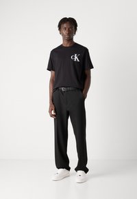 Svart kortermet t-skjorte med sølv "cK"-logo, kombinert med sorte bukser og hvite sneakers, som gir et uformelt og moderne utseende.