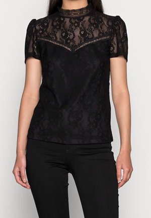 Woman wearing a black lace short-sleeve top with high neck and floral patterns, paired with black pants, standing against a plain backdrop.