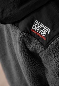 Black and gray fleece jacket with "SUPER DRY® Since 2003" embroidered in white and red on the upper chest area.