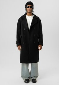 Man draagt een zwarte oversized dubbelgeknoopte jas, wit overhemd, lichtblauwe wijde jeans, zwarte schoenen, zwarte beanie, zonnebril en zilveren ketting.