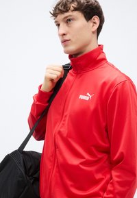 Κόκκινο μπουφάν με φερμουάρ κατασκευασμένο από λείο, ελαφρύ υλικό. Διαθέτει ψηλό γιακά και λευκό λογότυπο Puma. Μαύρη τσάντα τύπου duffel φοριέται στον ώμο.