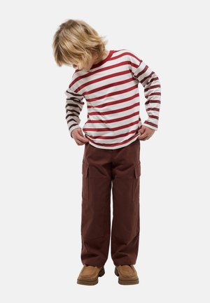 Enfant aux cheveux blonds portant un t-shirt à manches longues rayé rouge et blanc, un pantalon cargo marron et des chaussures beige, regardant vers le bas et ajustant sa chemise.