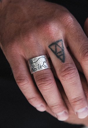 Bague large en argent avec un motif de lion en relief. La main présente un teint clair, avec un tatouage géométrique sur l'annulaire.