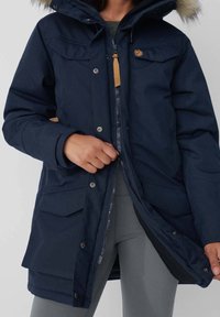 Geacă parka de marină cu glugă căptușită cu blană, închidere cu fermoar, buzunare frontale și accente din piele bej. Materialul pare durabil și are o textură netedă.