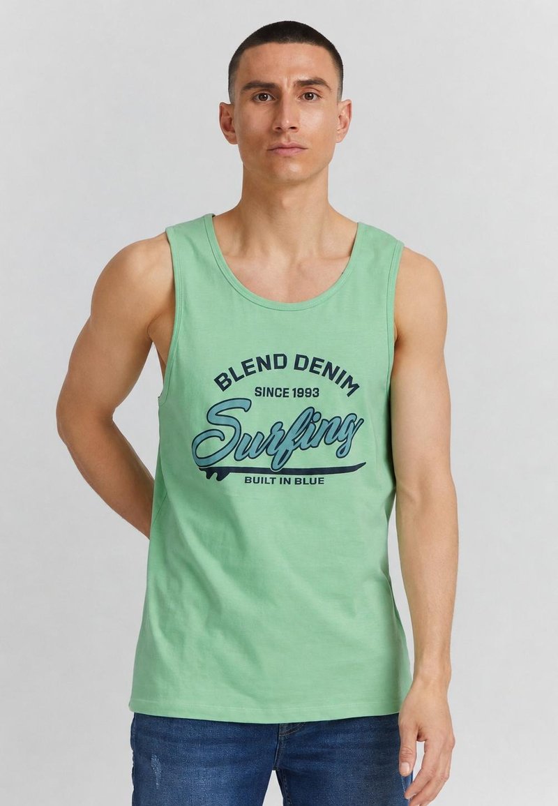 Homme portant un débardeur vert clair avec le texte "Blend Denim Surfing since 1993" et un jean bleu, debout devant un fond gris uni.