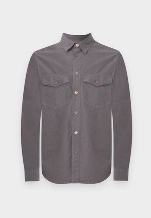 Férfi áll, kezei a zsebében, fekete nadrágot, fekete grafikás pólót, szürke gombos inget és fekete cipőt visel fehér háttér előtt.