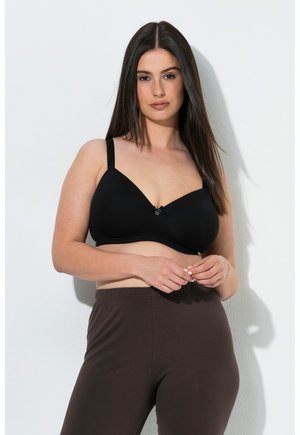 Donna con lunghi capelli scuri che indossa un reggiseno nero e pantaloni marrone scuro, in piedi contro un muro bianco con le mani leggermente intrecciate.