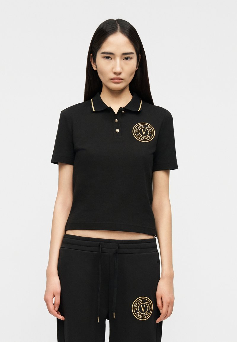 Γυναίκα που φοράει μαύρο πόλο με κοντά μανίκια και παντελόνι που ταιριάζει, με λογότυπα Versace Jeans Couture με χρυσές λεπτομέρειες στο στήθος και στον μηρό.