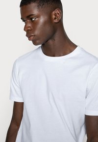 Weißes Baumwoll-T-Shirt mit Rundhalsausschnitt, kurzen Ärmeln und glatter Textur, getragen von einer Person, die leicht zur Seite blickt.