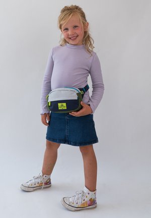 Sac banane gris clair et bleu marine avec des accents verts ; compartiments zippés ; porté sur un haut lilas à manches longues et une jupe en denim.
