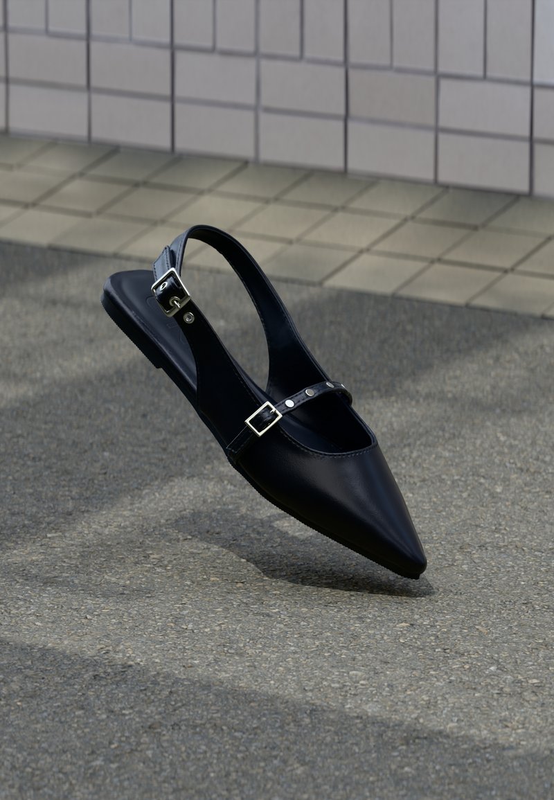 Μαύρο φλατ παπούτσι slingback με μυτερή μύτη και διπλές λουράκια με αγκράφες, τοποθετημένο σε γκρι άσφαλτο κοντά σε τοίχο με πλακάκια.