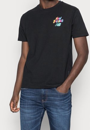 Schwarzes Kurzarm-T-Shirt aus Baumwolle mit einem bunten "New Balance"-Logo auf der linken Brust. Kombiniert mit blauen Denim-Jeans.