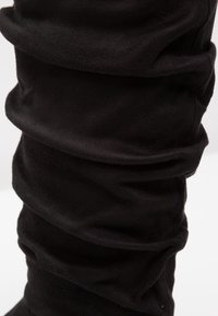 Gros plan sur une botte haute molletonnée en daim noir, avec des plis texturés doux et un léger éclat, sur un fond clair uni.