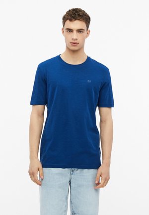 Jonge man met kort krullend haar draagt een effen blauw t-shirt met korte mouwen en lichtblauwe spijkerbroek, staand tegen een witte achtergrond.