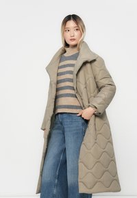 Vrouw in een lange beige gewatteerde jas over een gestreepte beige en grijze sweater met wijde spijkerbroeken, staand tegen een witte achtergrond.