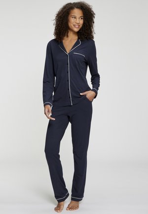 Vrouw die blootsvoets staat en een donkerblauw pyjamapak met lange mouwen en witte bies draagt, glimlacht en kijkt opzij met één hand in de zak.