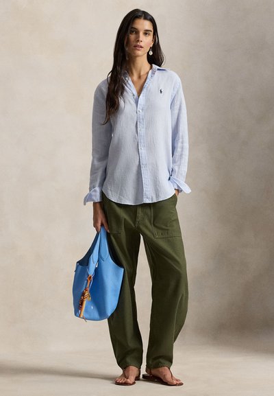 Γυναίκα με ανοιχτό γαλάζιο πουκάμισο με κουμπιά και χακί παντελόνι, κρατώντας μεγάλη μπλε τσάντα, με γυμνά πόδια και όρθια μπροστά σε απλό φόντο.