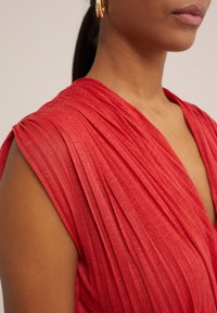 Röd plisserad klänning med djup V-urringning och korta ärmar. Tyget är strukturerat, med intrikata linjer och draperingar. Enkla designinslag.