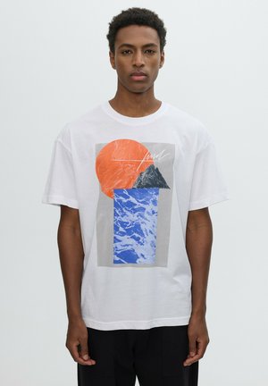 Hombre que lleva una camiseta blanca con un diseño abstracto que muestra un círculo naranja, una cima de montaña negra y un patrón de agua azul en la parte delantera.