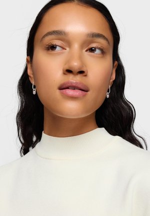 Młoda kobieta o długich ciemnych włosach, nosząca srebrne kolczyki wiszące i biały sweter z wysokim kołnierzem, spoglądająca lekko na bok na tle jednolitego tła.