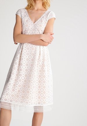 Frau trägt ein knieumspieltes weißes Spitzenkleid mit kurzen Ärmeln und V-Ausschnitt, steht mit verschränkten Armen vor einem schlichten Hintergrund.