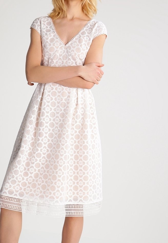 Femme portant une robe blanche en dentelle longueur genoux, à manches courtes et col en V, debout les bras croisés devant un fond uni.
