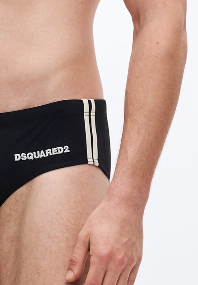 Schwarze Badehose mit beigen Seitenstreifen, versehen mit dem gestickten "DSQUARED2"-Logo auf der linken Hüfte. Der Stoff wirkt glatt und eng anliegend.