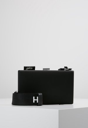 Pochette rectangulaire noire avec ferrures argentées et une bandoulière noire détachable ornée d'un "H" blanc, posée sur une surface blanche unie.
