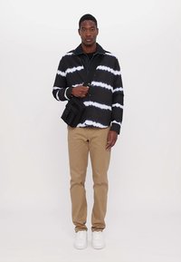 Sort og hvid tie-dye jakke, knap lukket, båret over en sort skjorte. Tan bukser og hvide sneakers fuldender outfittet. Sort skuldertaske inkluderet.