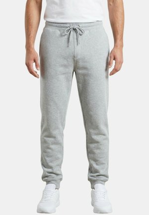 Homme portant un pantalon de survêtement gris clair avec taille à cordon, associé à des baskets blanches et un t-shirt blanc, debout devant un fond uni.