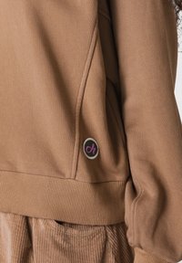 Felpa marrone con polsi a costine. Presenta una toppa rotonda con un logo rosa contornato di bianco, situata vicino all'orlo. Tessuto morbido.