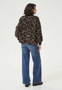 Maglione marrone con stampa leopardata nera, maniche a costina e vestibilità ampia, abbinato a jeans blu a gamba larga e calzature nere.