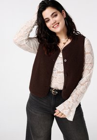 Bruin fleecestukken met afgeronde zoom, voorzien van knopen aan de voorkant, gedragen over een witte kanten top met uitlopende mouwen, gecombineerd met donkere jeans.