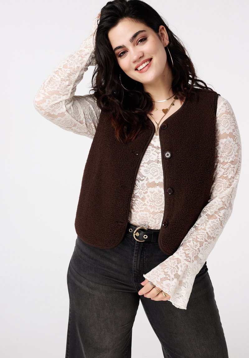 Bruin fleecestukken met afgeronde zoom, voorzien van knopen aan de voorkant, gedragen over een witte kanten top met uitlopende mouwen, gecombineerd met donkere jeans.